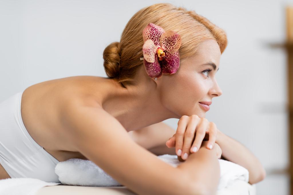 vue latérale de la femme blonde heureuse avec orchidée dans les cheveux couchés sur la table de massage  - Photo, image