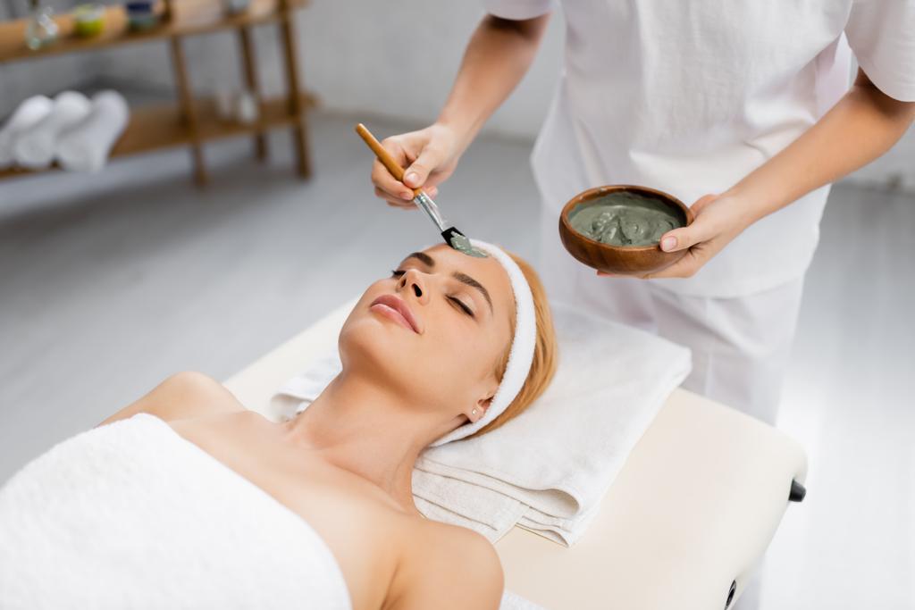esteticista aplicación de máscara de arcilla en la cara de la mujer acostada en la mesa de masaje - Foto, Imagen