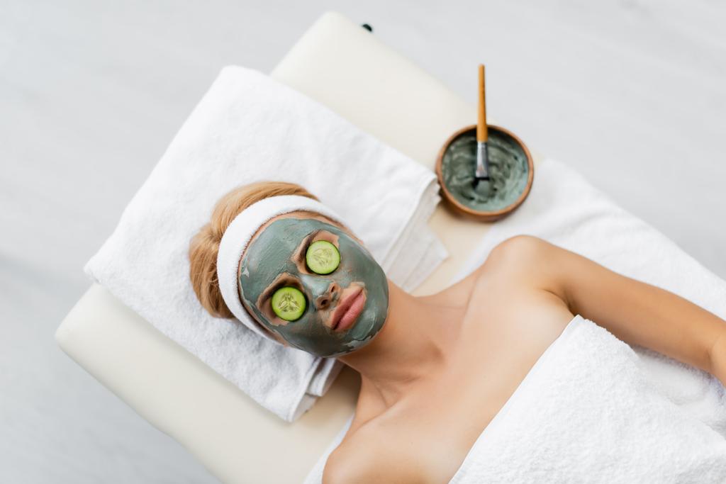 vista superior da mulher com máscara de barro e fatias de pepino em olhos deitados na mesa de massagem - Foto, Imagem