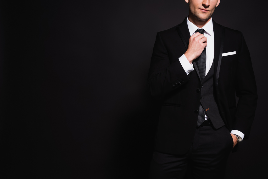 частковий вид елегантного чоловіка торкається краватки, стоячи з рукою в кишені ізольовані на чорному
 - Фото, зображення