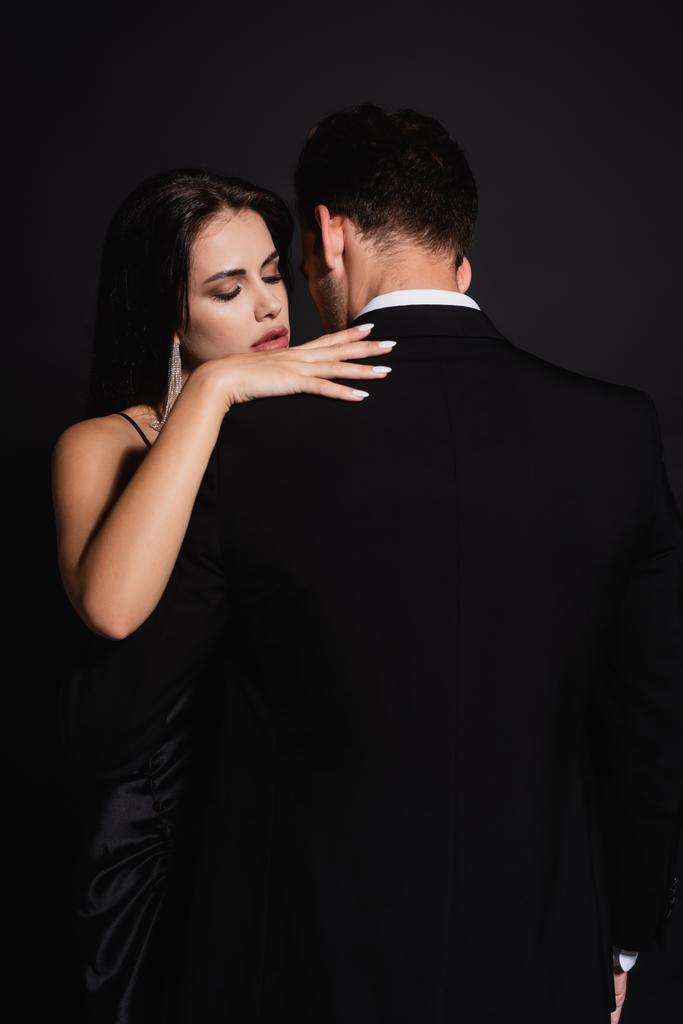 back view of man in suit near elegant woman hugging him isolated on black - Photo, Image