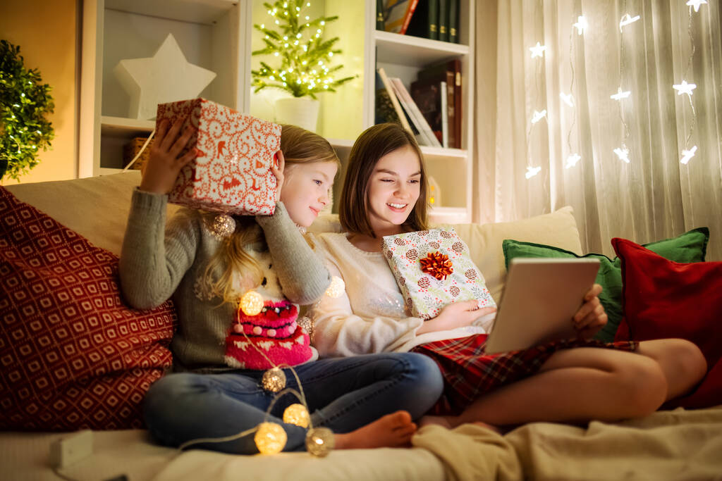 Noel 'de evde sıcak ve rahat oturma odasında tablet kullanan iki kız kardeş. Noel arifesinde ailenin online video görüşmesi var. Aile ve çocuklarla evde kış akşamı. - Fotoğraf, Görsel
