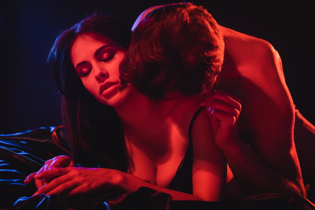 jeune homme embrasser femme passionnée couché sur la literie de soie dans l'éclairage rouge sur fond noir - Photo, image