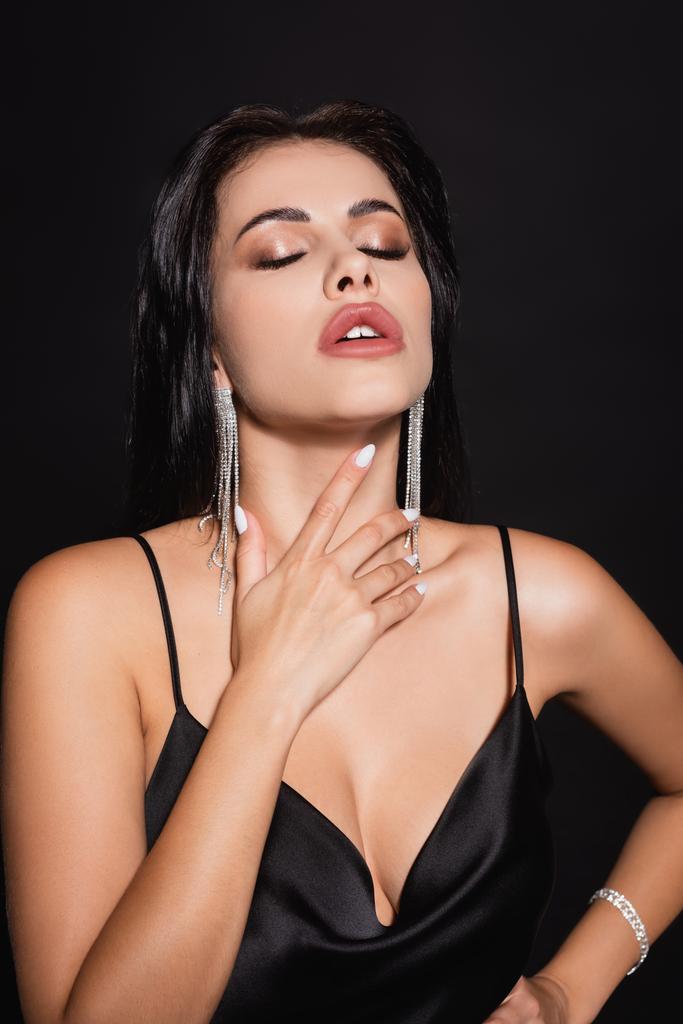 sexy Frau im Seidenkleid und Ohrringe berühren Hals, während sie mit geschlossenen Augen isoliert auf schwarz posiert - Foto, Bild