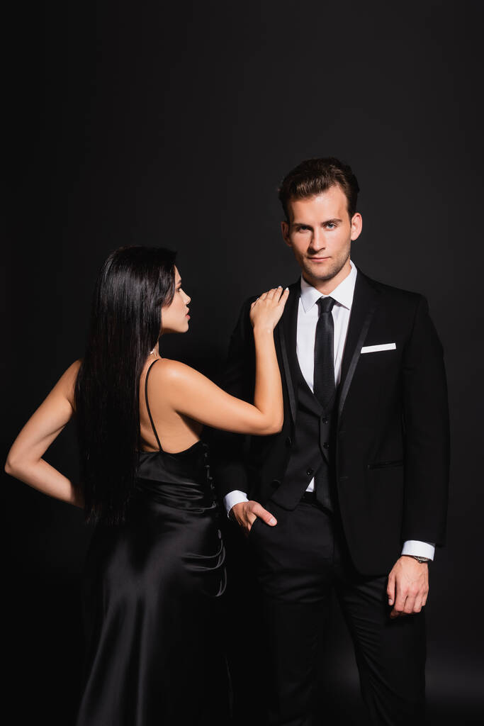 brunette vrouw in zijde jurk aanraken schouder van elegante man staande met de hand in zak op zwart - Foto, afbeelding
