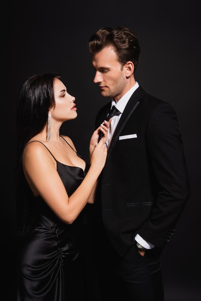 brunette vrouw verleiden man in elegante pak en aanraken van zijn das geïsoleerd op zwart - Foto, afbeelding