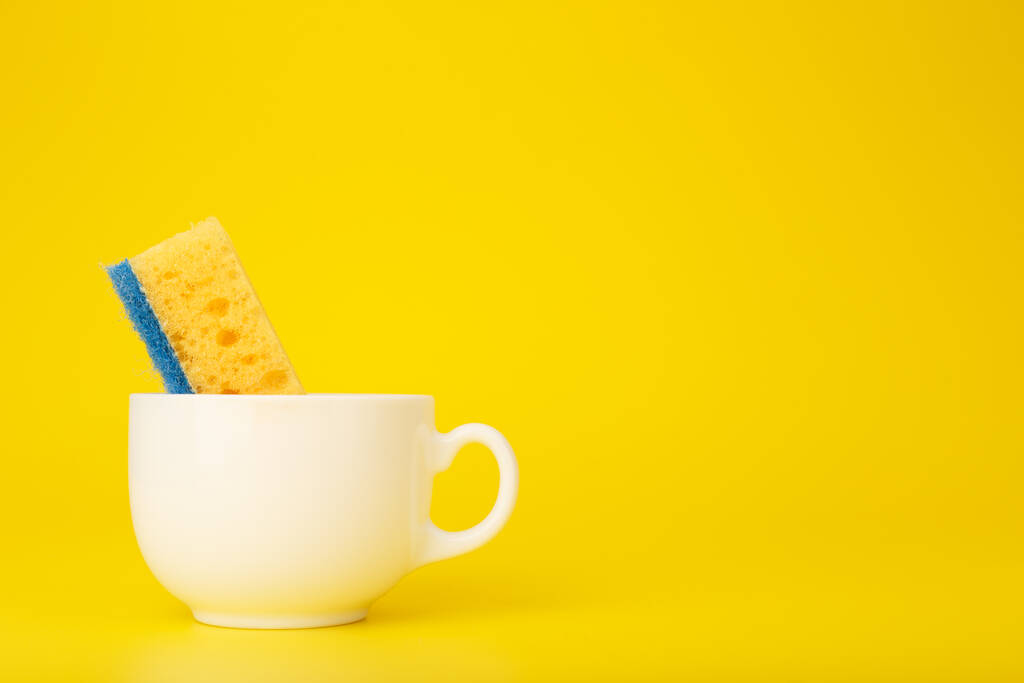 Concept de lave-vaisselle créatif et minimal. Composition simple avec éponge de cuisine de nettoyage jaune en tasse en céramique blanche sur fond jaune avec espace de copie.  - Photo, image