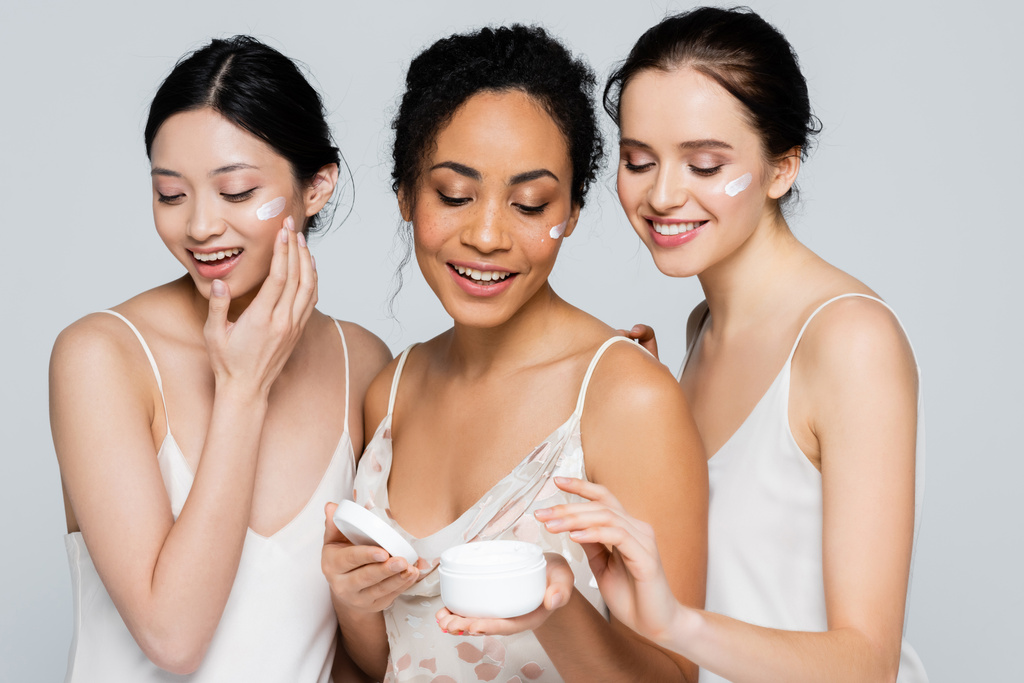 Felices mujeres multiculturales aplicando crema cosmética en rostros aislados en gris  - Foto, imagen