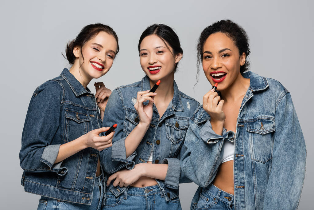 Pretty multiethnic women in denim jackets holding red lipsticks and looking at camera isolated on grey  - Photo, Image