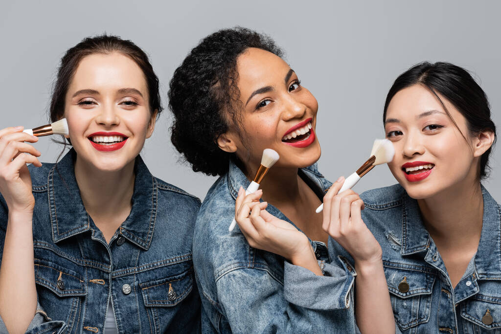 Kırmızı dudakları olan neşeli ırklar arası kadınlar gri yüzlerin yakınında kozmetik fırçalar tutuyorlar.  - Fotoğraf, Görsel