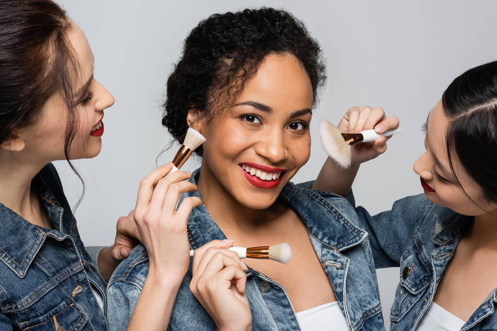 Smiling african american woman in denim jacket standing near interracial friends with cosmetic brushes isolated on grey  - Photo, Image