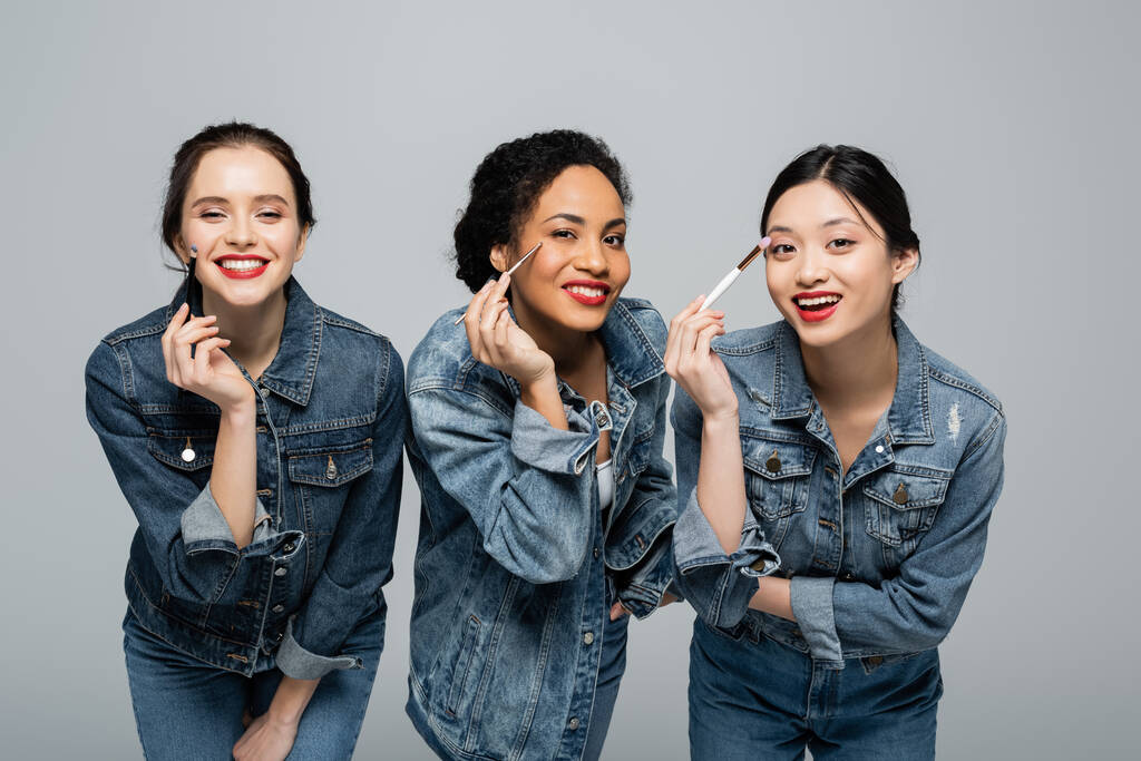 Lächelnde multiethnische Frauen in Jeansjacken halten Kosmetikbürsten in der Hand und blicken vereinzelt in die Kamera auf grau  - Foto, Bild