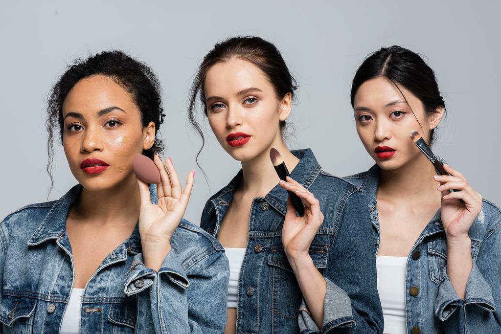 Young multiethnic women in denim jackets holding cosmetic brushes and beauty blender isolated on grey  - Photo, Image