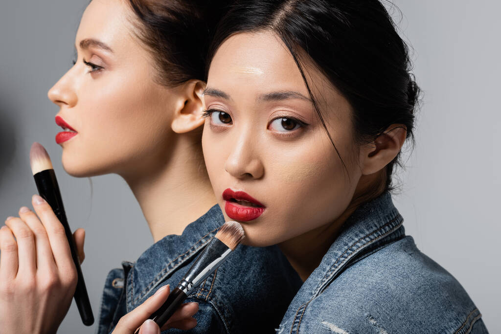 Aasian nainen punaiset huulet ja kasvot säätiö tilalla kosmeettinen harja lähellä ystävä harmaalla pohjalla  - Valokuva, kuva