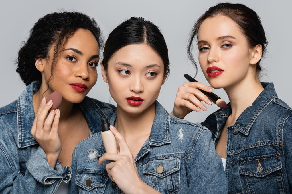 Mujeres jóvenes interraciales sosteniendo cepillo cosmético, fundación de la cara y licuadora de belleza aislado en gris  - Foto, imagen