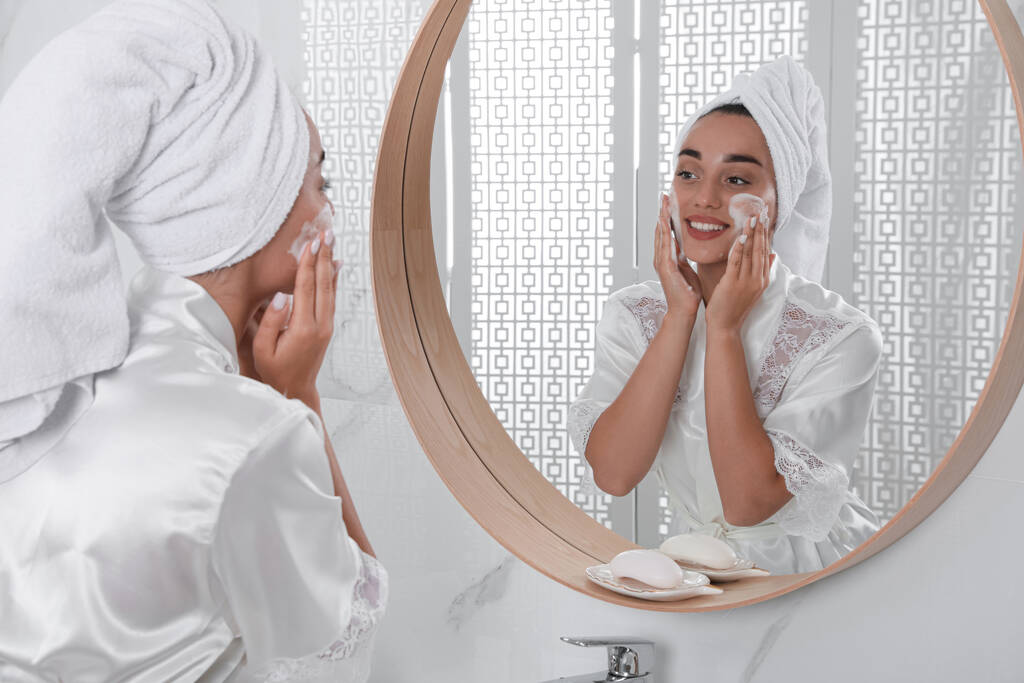 Hermosa mujer joven aplicando espuma limpiadora en la cara cerca del espejo en el baño. Cuidado de la piel cosmética - Foto, Imagen