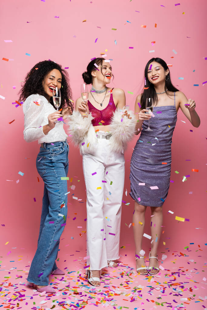 Cheerful interracial women with champagne celebrating new year under confetti on pink background - Photo, Image
