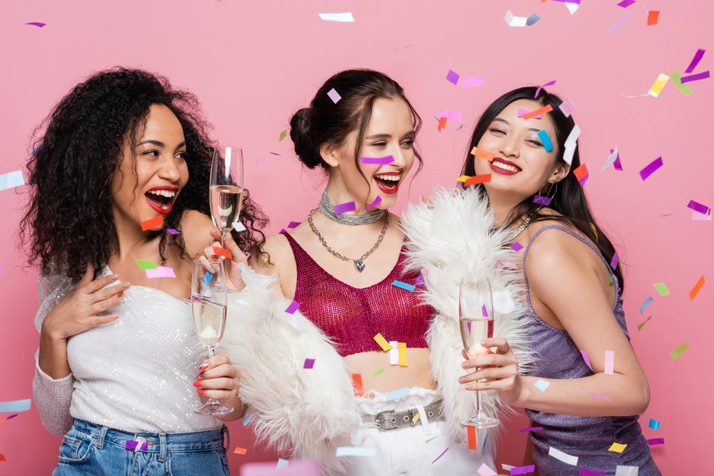 Élégantes femmes multiethniques tenant des verres de champagne sous confettis sur fond rose - Photo, image