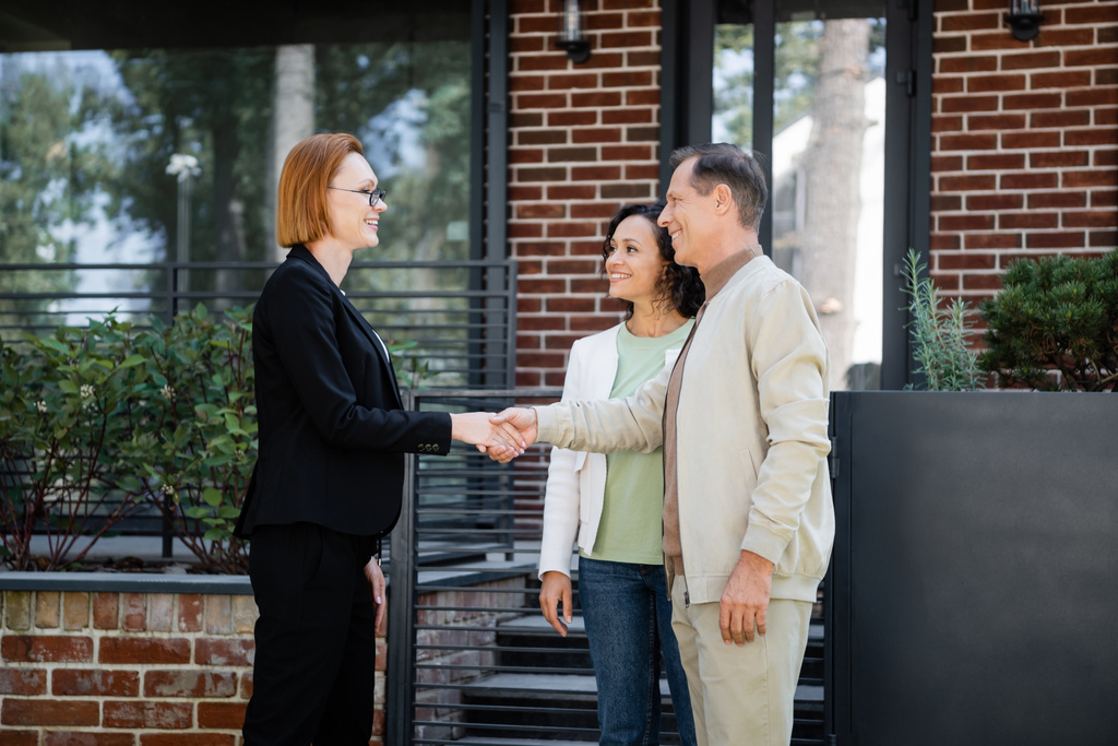 happy realtor in glasses shaking hands with man standing near african american wife and modern house - Photo, Image