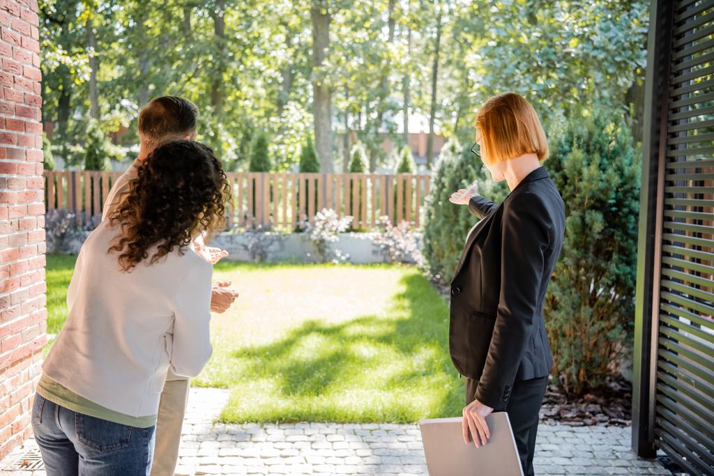 back view of redhead realtor in glasses pointing at lawn to couple  - Photo, Image