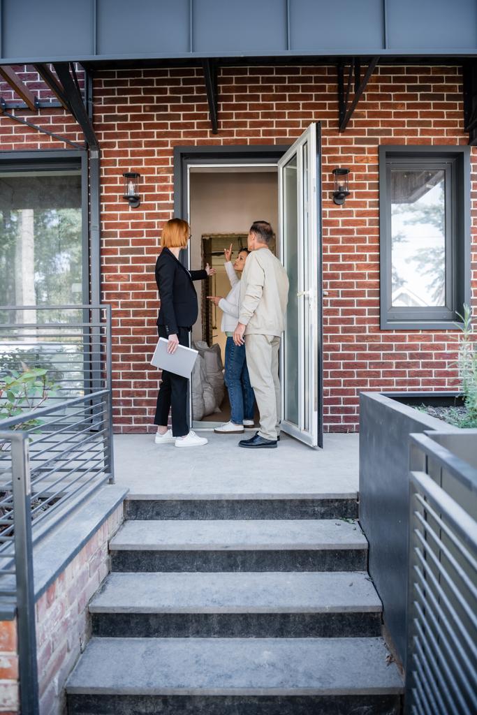 rousse agent immobilier dans des lunettes pointant du doigt à la maison tout en parlant avec le couple interracial - Photo, image
