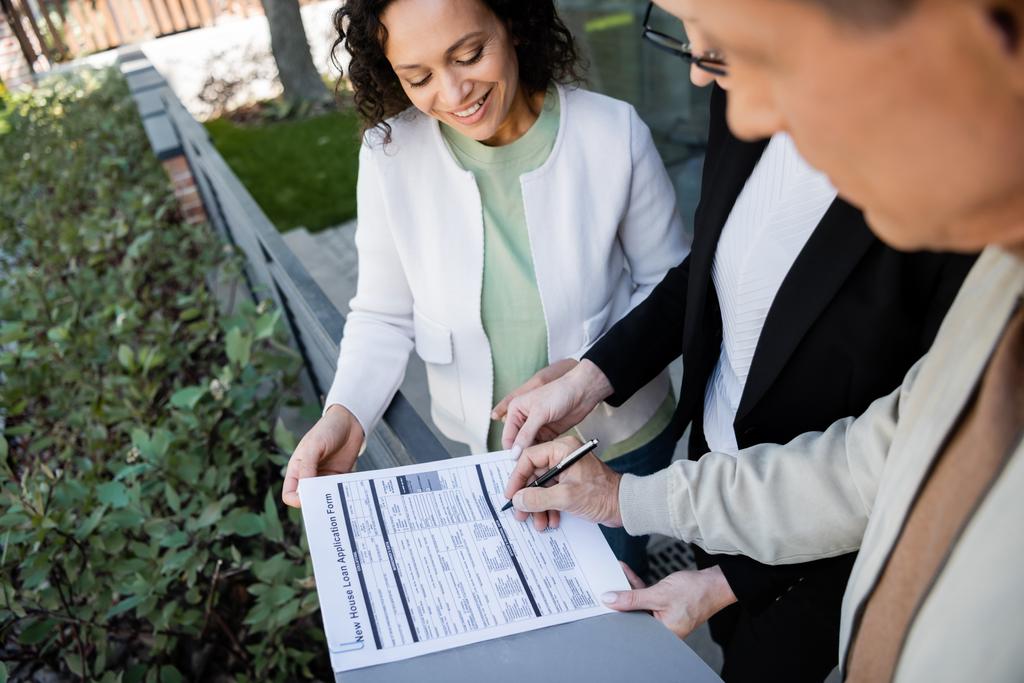 hombre borroso firma solicitud de préstamo cerca de esposa afroamericana feliz y agente inmobiliario  - Foto, imagen