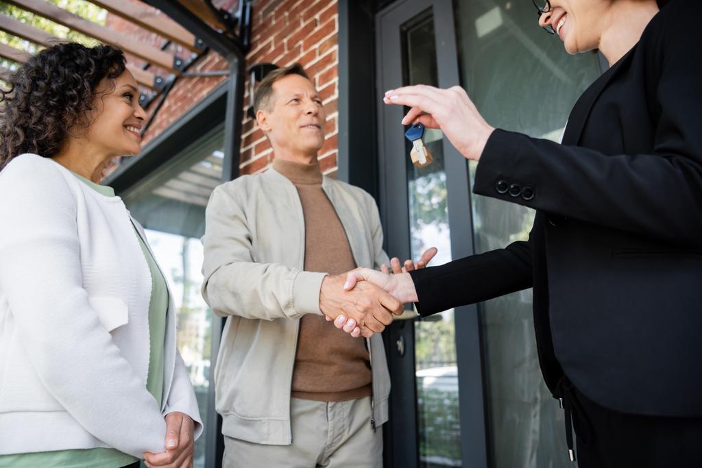low angle view of happy realtor holding key while shaking hands with cheerful man near african american wife - Photo, Image