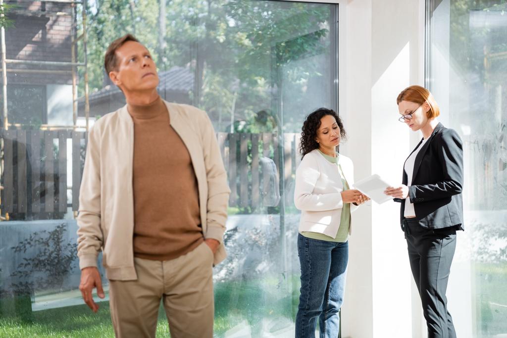uomo sfocato in piedi con la mano in tasca vicino afroamericano donna parlando con redhead agente immobiliare mentre guardando tablet - Foto, immagini