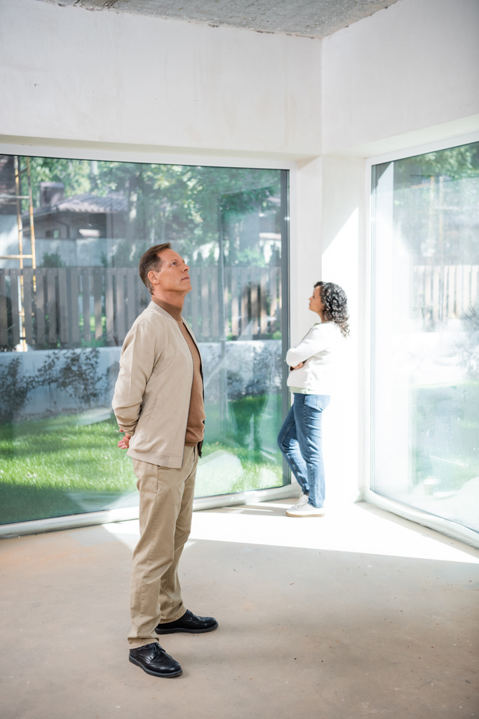 uomo guardando soffitto vicino afro-americano moglie in piedi vicino a finestre panoramiche in nuova casa - Foto, immagini