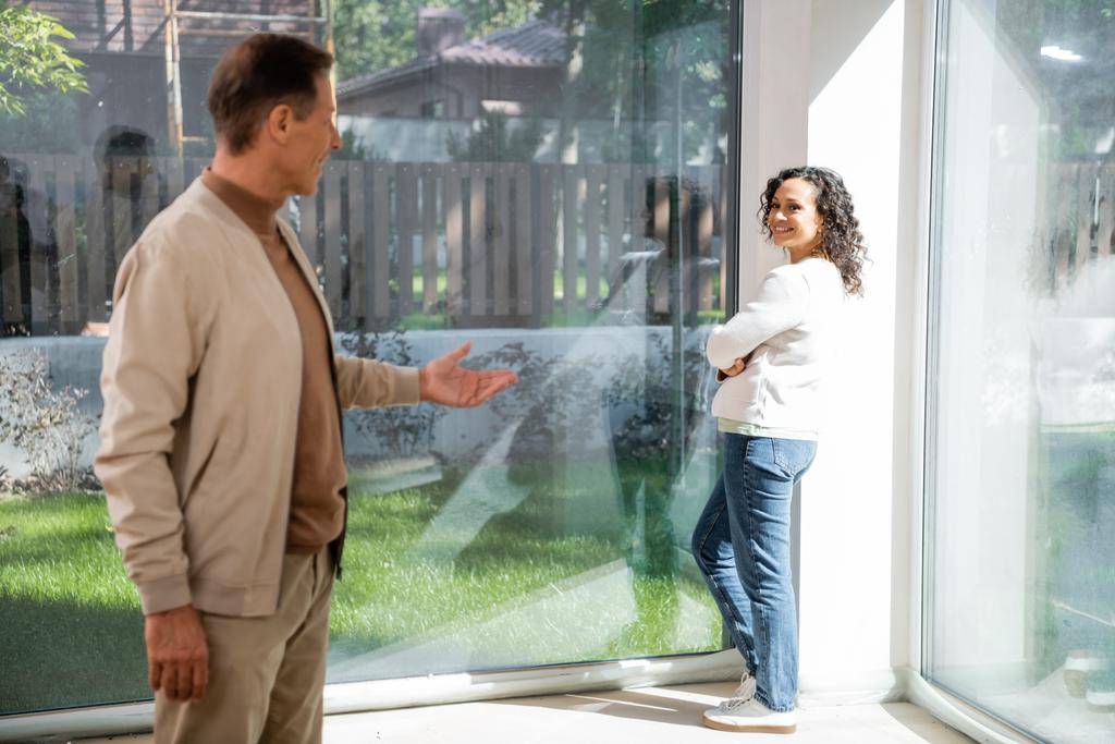 mujer afroamericana de pie cerca de ventanas panorámicas mientras mira marido borroso en casa nueva - Foto, Imagen