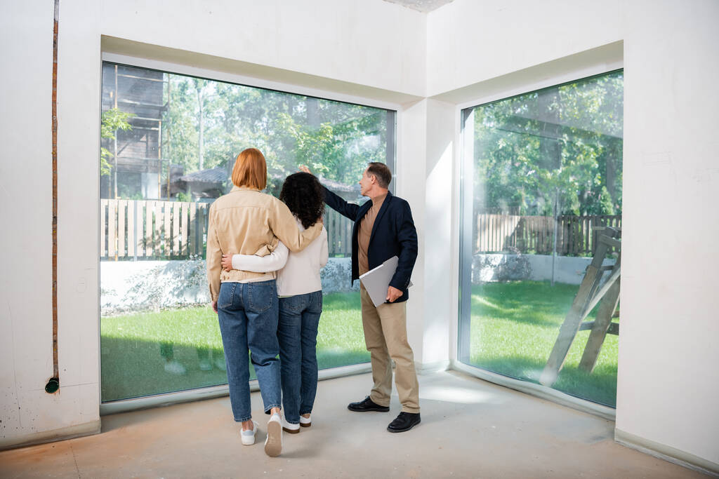 agent immobilier d'âge moyen tenant dossier et pointant vers la fenêtre panoramique près de couple lesbien interracial à l'intérieur de la nouvelle maison - Photo, image