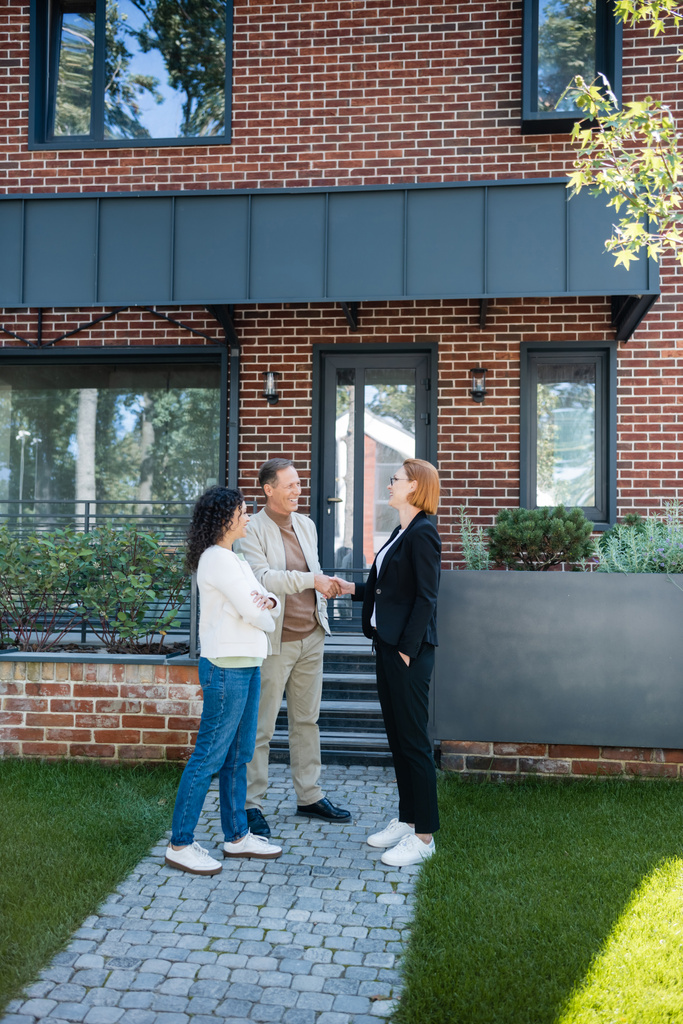 alegre agente inmobiliario en gafas estrechando la mano con sonriente hombre de pie cerca de la esposa afroamericana y la casa moderna - Foto, imagen