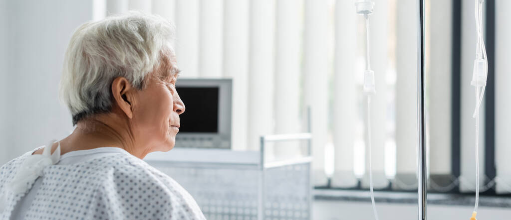 Вид збоку на старшого пацієнта в сукні, який дивиться на внутрішньовенну терапію в лікарняному відділенні, банер
  - Фото, зображення