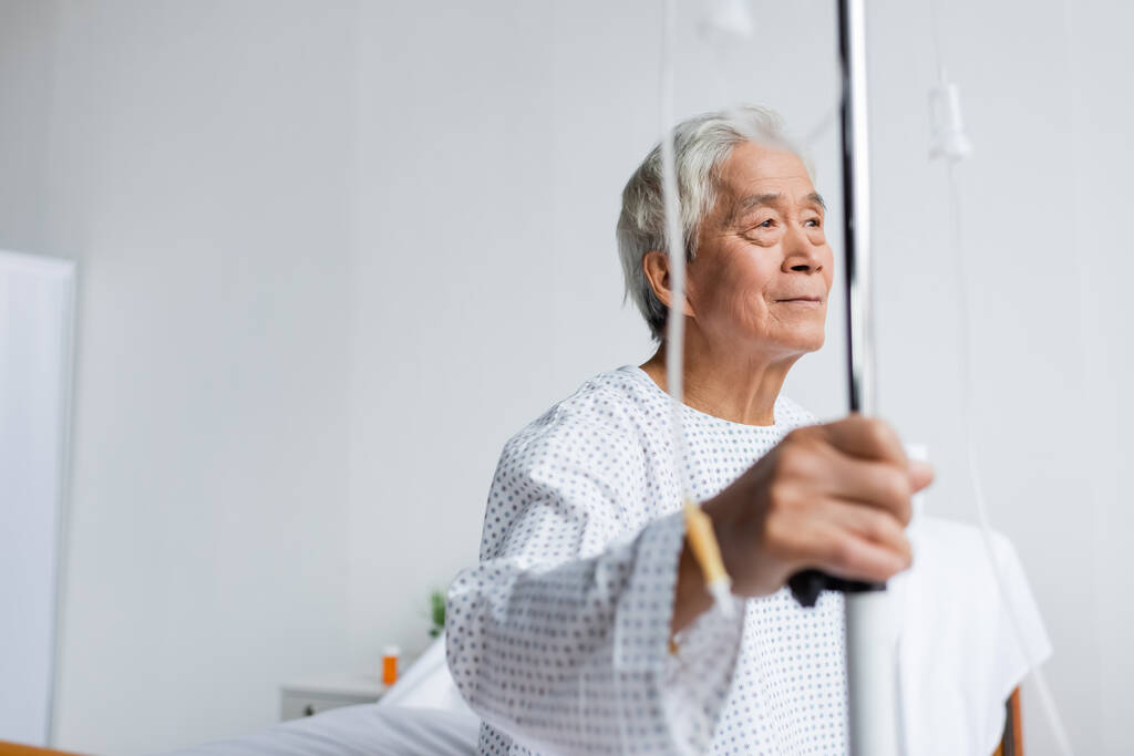 Літній азіатський пацієнт тримає розмиту підставку з внутрішньовенною терапією в лікарняному відділенні
  - Фото, зображення