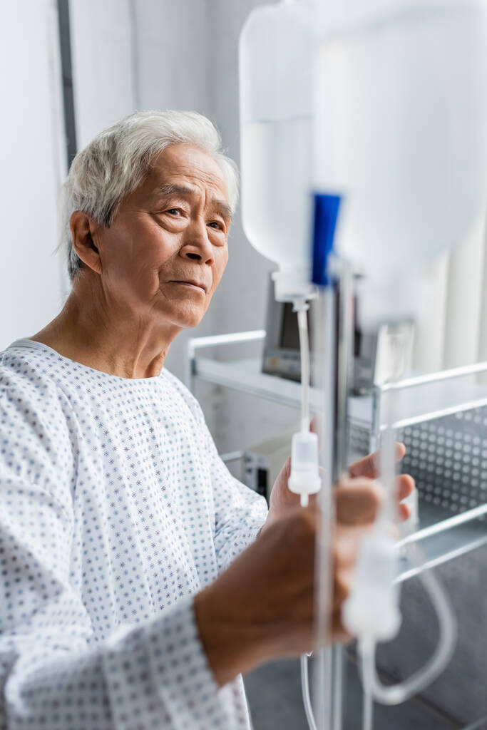 Asian patient in gown looking at blurred intravenous therapy in hospital ward - Photo, Image