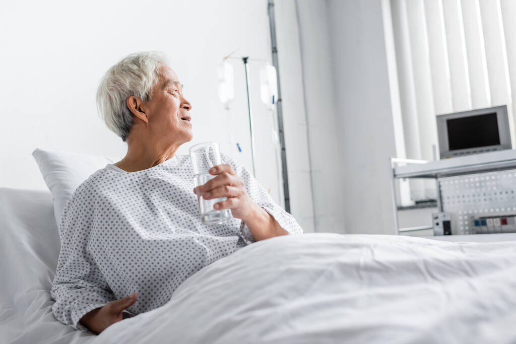 Asyalı yaşlı hasta hastane yatağında bir bardak su tutuyor.  - Fotoğraf, Görsel