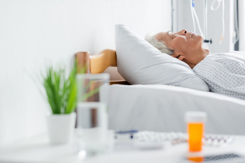 Vista lateral de paciente asiática mayor durmiendo en la cama cerca de agua borrosa y pastillas en la clínica  - Foto, imagen