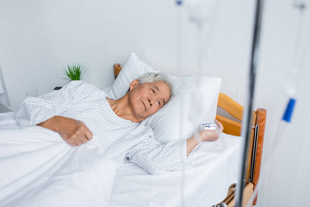 Senior asian man with pulse oximeter lying near blurred intravenous therapy on bed in hospital  - Photo, Image