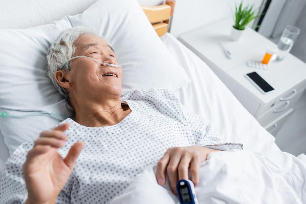 Hoge hoek uitzicht van lachende Aziatische patiënt met pulsoximeter en nasale canule liggend op bed in kliniek  - Foto, afbeelding
