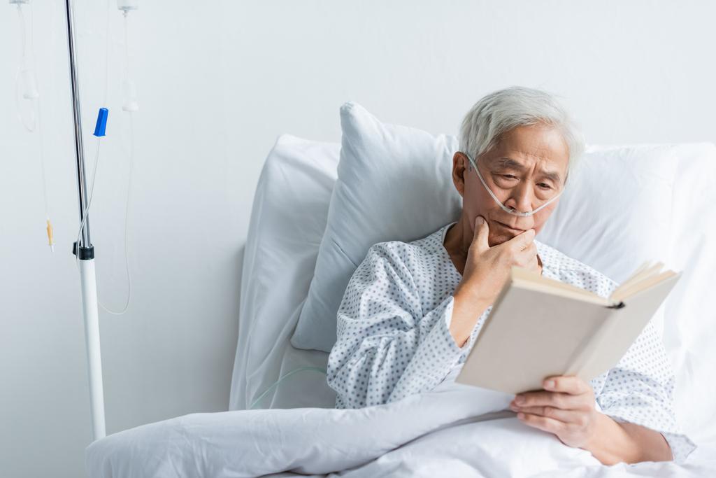 Oudere Aziatische patiënt met nasale canule leesboek op bed in kliniek  - Foto, afbeelding