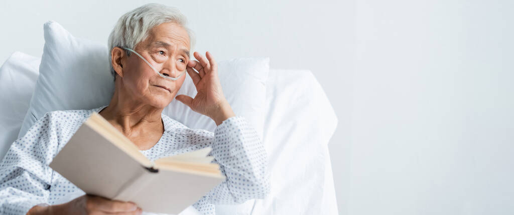 Ηλικιωμένος Ασιάτης ασθενής με ρινική κάνουλα κρατώντας βιβλίο στο κρεβάτι του νοσοκομείου, πανό  - Φωτογραφία, εικόνα