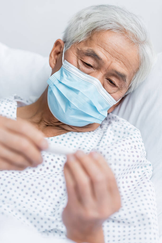 Tıbbi maskeli kıdemli Asyalı hasta yatağında bulanık termometre tutuyor.  - Fotoğraf, Görsel