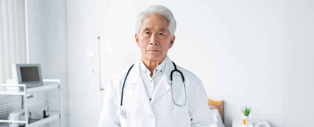 Старший азиатский врач смотрит в камеру в клинике, баннер  - Фото, изображение