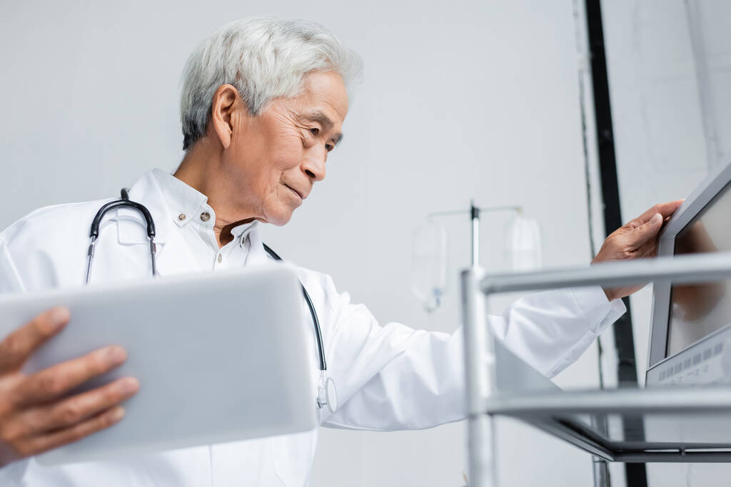 Älterer asiatischer Arzt hält verschwommenes digitales Tablet in der Nähe medizinischer Geräte in Klinik  - Foto, Bild