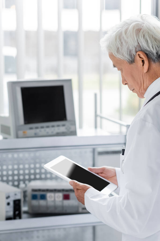 Idős ázsiai orvos digitális tablettát tart a kórházi homályos orvosi berendezések közelében   - Fotó, kép