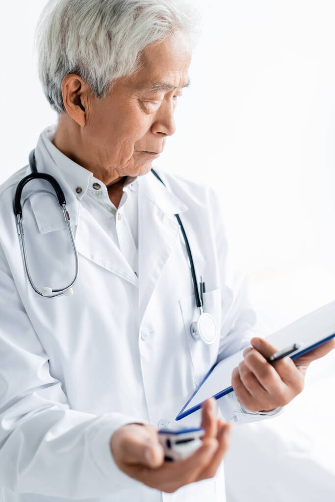 Ασιάτης γιατρός κοιτάζοντας πρόχειρο και κρατώντας θολή παλμικό οξύμετρο στην κλινική  - Φωτογραφία, εικόνα