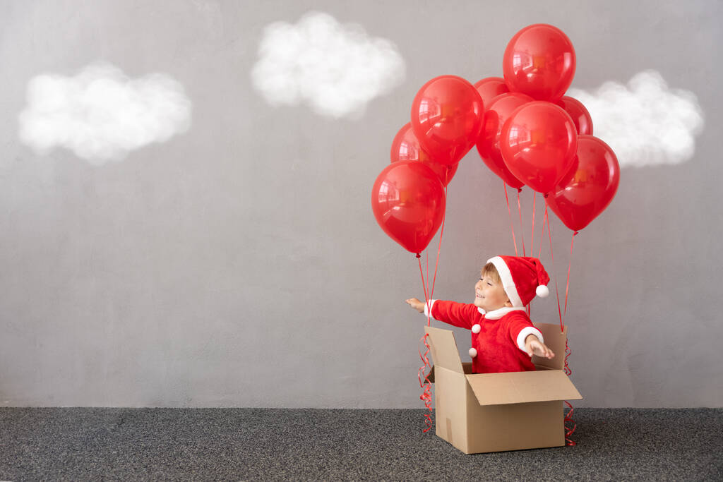 Happy child wearing Christmas costume. Kid sitting in cardboard box with red balloons. Funny child playing. Kid flying ahead. Xmas gift box concept - Photo, Image