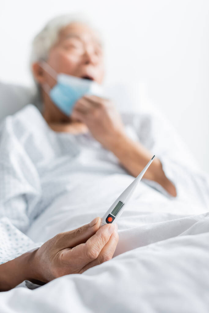 Ηλεκτρονικό θερμόμετρο στο χέρι σε θολή ηλικιωμένη ασθενή στο θάλαμο του νοσοκομείου  - Φωτογραφία, εικόνα