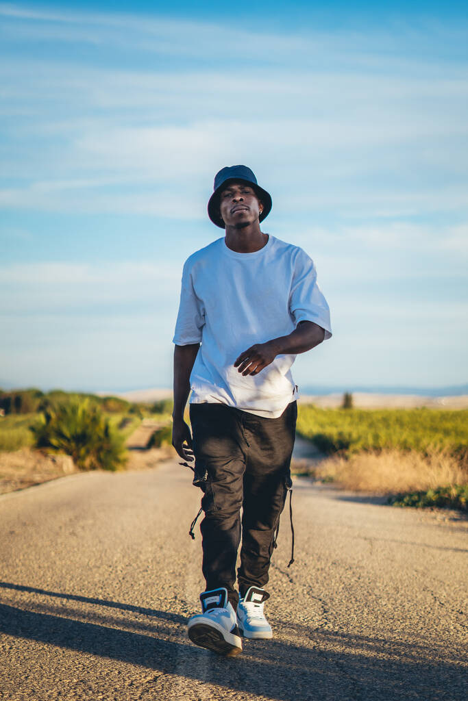 Un disparo de cuerpo completo de un joven guapo negro con un sombrero de cubo caminando por la carretera, puesta de sol - Foto, imagen