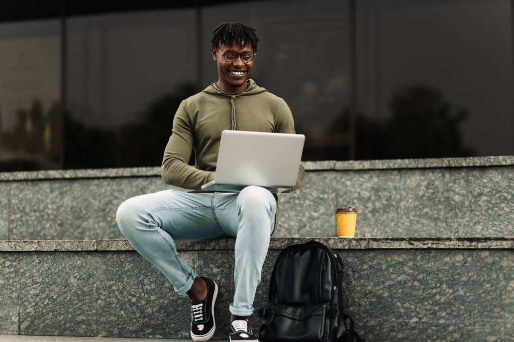 Улыбающийся африканский мужчина, используя ноутбук, сидя на городской лестнице, делая заметки, наблюдая вебинар, занимаясь онлайн-уроком, мотивированный позитивный студент учится в Интернете, глядя на экран ноутбука, на открытом воздухе - Фото, изображение